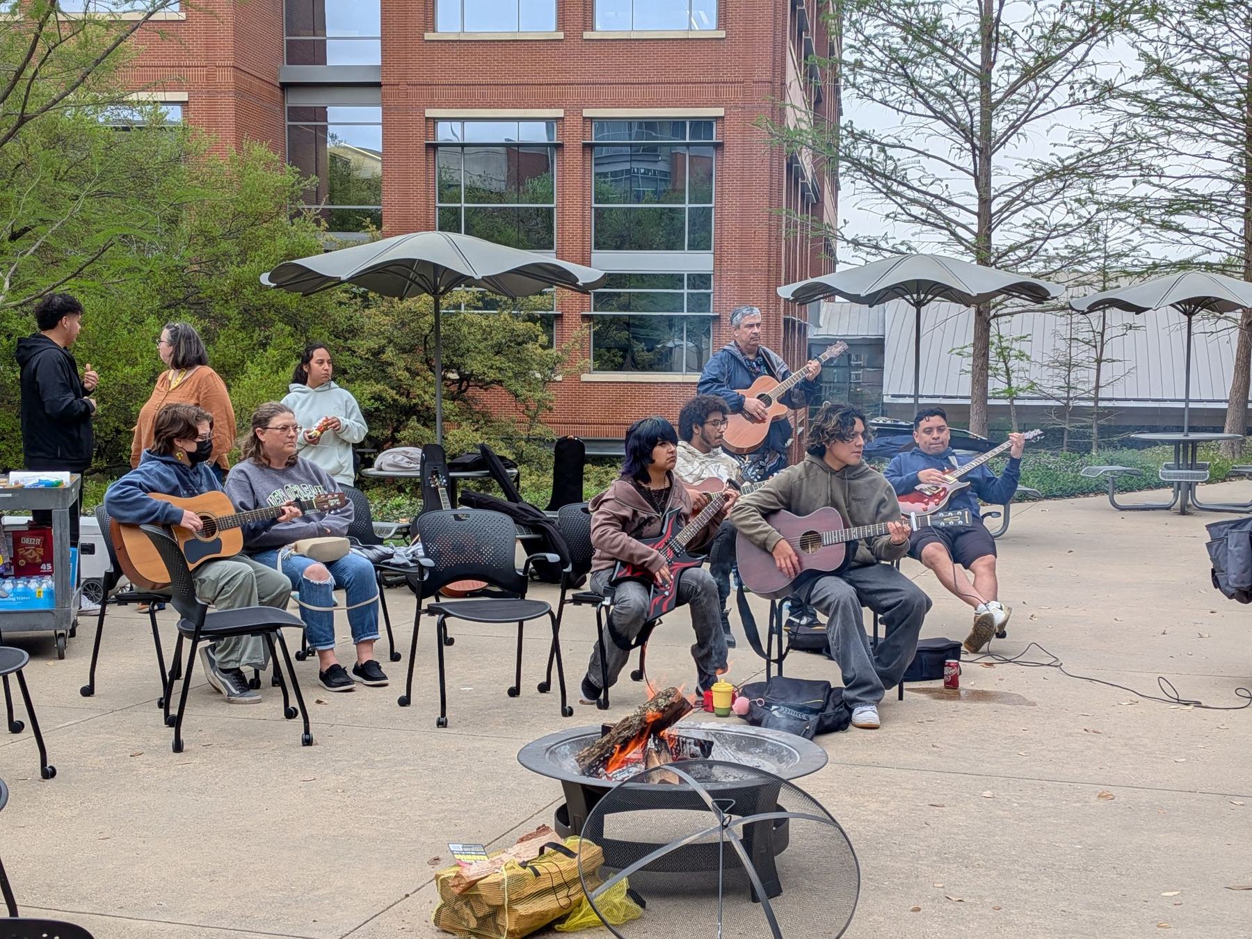 Students seated in a circle around a fire pit on campus, playing acoustic and electric guitars together