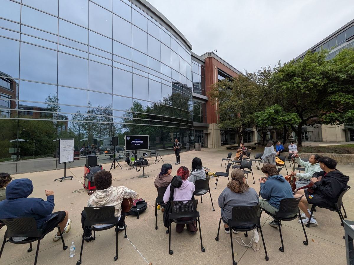 Wide view of the guitar club gathered in a circle of chairs outside a glass campus building at dusk