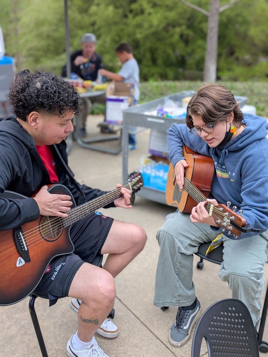 Two students sitting side by side, one showing the other a chord shape on an acoustic guitar