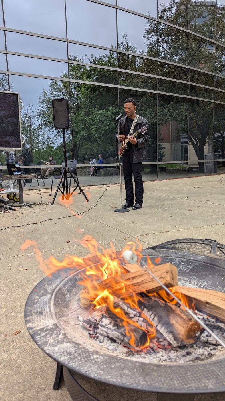 A performer singing into a microphone beside a burning fire pit at the Campfire Strum Along